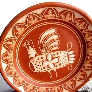 May include: A round terracotta plate featuring a central white graphic illustration of a stylized bird with a patterned body and a four-petaled flower design. The rim is decorated with a repeating white leaf pattern against the reddish-brown clay.