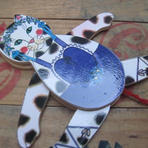 May include: A whimsical wooden cat toy with a white face, black spots, and a blue heart-shaped body. The cat has a floral crown and is painted with pink cheeks and green eyes. The toy is connected with red string.