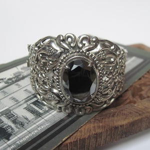 May include: A silver-toned cuff bracelet with an oval, dark gemstone center. The bracelet features ornate, detailed scrollwork and is set on a vintage photograph and a wooden surface. The bracelet is a statement piece.