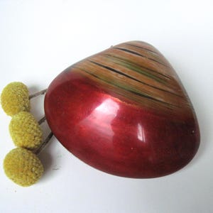 May include: A glossy, heart-shaped brooch with a gradient of red to brown hues. The top section features brown and green stripes. Three yellow, spherical flower buds are positioned to the left of the brooch.