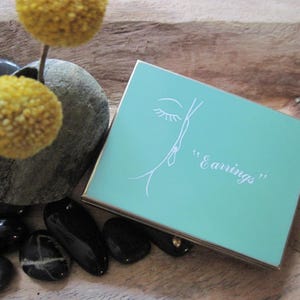 May include: A vintage teal jewelry box with gold trim, labeled "Earrings" in script, features a stylized face illustration. The box rests on a wooden surface with black stones and yellow floral accents.