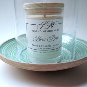 May include: A soy wax candle in a glass jar with a wooden lid, labeled "Island Memories SF Bora Bora." The candle sits on a round, wooden tray with a turquoise and white spiral design, inside a clear glass cylinder.