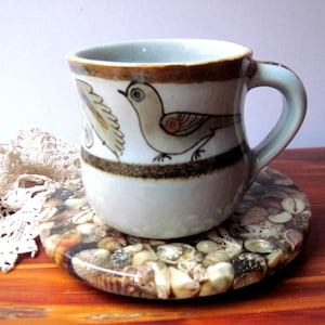 May include: A ceramic mug with a light gray glaze and a hand-painted brown bird motif, featuring a dark brown horizontal band near the rim and base. The mug rests on a circular coaster embedded with a dense collection of small, multi-colored seashells.
