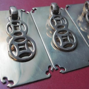 May include: Three rectangular brass pendants with a decorative, openwork design. Each pendant features a loop at the top and a stylized, repeating pattern. The pendants are displayed on a red surface.