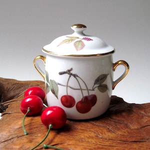 May include: A white ceramic sugar bowl with a lid, gold trim, and handles. The bowl features a cherry branch design with red cherries and green leaves. Three red cherries with green stems are placed in front of the bowl.