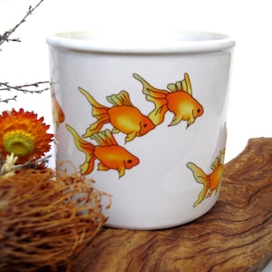 May include: A white ceramic mug featuring a repeating graphic print of bright orange goldfish with flowing fins. The mug has a smooth, cylindrical body and a simple rim, showcasing a clean design that highlights the vibrant aquatic illustrations.