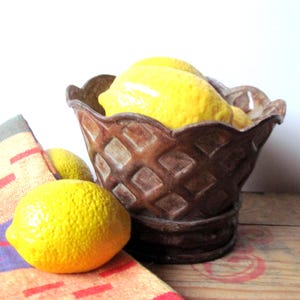Puede incluir: Una cesta de cerámica marrón con un patrón de diamantes, llena de limones amarillos brillantes. Un limón descansa fuera de la cesta sobre una toalla estampada colorida. La cesta está sobre una superficie de madera.