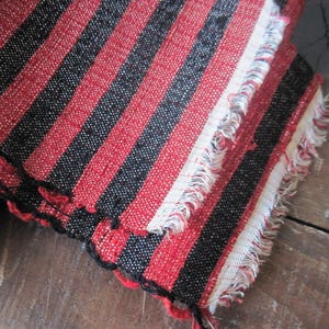 May include: Red and black striped fabric with a fringed edge. The fabric has a woven texture and is folded over on itself.