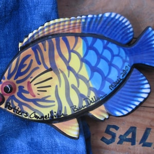 May include: A ceramic fish figurine with a vibrant design. The fish features blue, yellow, and orange patterns, with the text "Potters Angel Fish" and "Maui 2001" visible. The figurine is placed on a wooden surface.