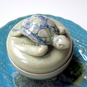 May include: A ceramic trinket box with a turtle figurine on top. The turtle has a blue and white shell, and the box is light green. The box is sitting on a blue, patterned plate.