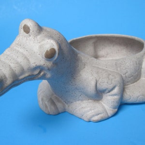 May include: A ceramic planter shaped like a gray alligator with a speckled texture. The alligator has large, round eyes and a wide open mouth. The planter has a shallow, round opening on the back for planting.