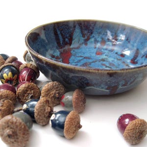 May include: A blue and red glazed ceramic bowl with a brown rim sits next to several decorative acorn ornaments. The acorns have brown caps and painted bases in various colors, including blue, red, and yellow.