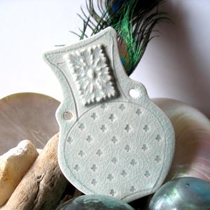 May include: A pale green ceramic decorative piece with a floral design and a cracked glaze. The piece has a unique shape with a handle and is surrounded by natural elements like shells and driftwood.