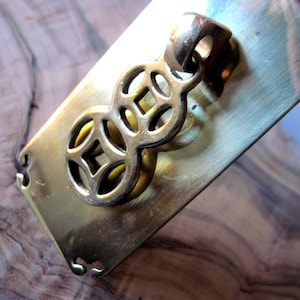 May include: A brass drawer pull with an ornate, openwork design. The rectangular backplate has decorative cutouts at the corners. The pull features a stylized, interlocking figure-eight shape with a central square element. The hardware has a warm, aged finish.
