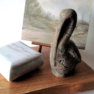 May include: A marble jewelry box with a lid sits on a wooden surface next to a weathered, abstract sculpture. A small painting on a stand is in the background. The sculpture is a dark, textured material.