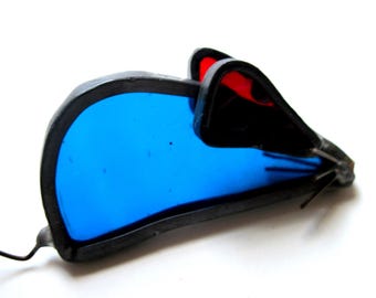 May include: A stained glass mouse ornament featuring a vibrant blue body, red inner ears, and black metal solder lines outlining the shape. The piece includes thin metal wire whiskers extending from the nose and a curved wire tail at the rear.