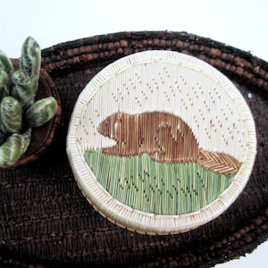 May include: A round, handcrafted decorative piece featuring a beaver design. The beaver is depicted in brown, resting on green grass, against a cream-colored background. The piece is surrounded by a woven border and rests on a dark brown woven tray.