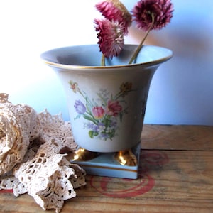 May include: A light blue porcelain vase with a flared rim and gold trim, featuring a floral bouquet print on the front. The vase stands on a square base with four gold-colored feet and contains three dried pink flowers extending from the top.