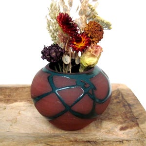 May include: A small, round, terracotta-colored ceramic vase with a teal geometric design. The vase holds a bouquet of dried flowers in shades of red, yellow, and beige. The vase sits on a wooden surface.