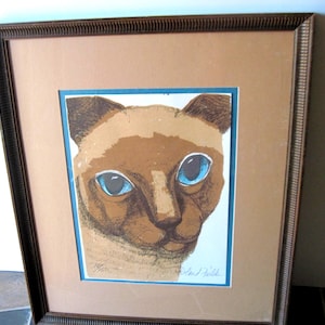 May include: Framed print of a Siamese cat's face. The cat has brown fur with striking blue eyes. The artwork is set against a light tan mat and framed in a dark brown, textured frame. The print is numbered 19/50.