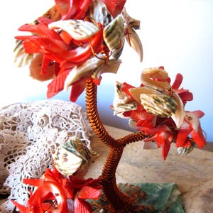 May include: A decorative tree sculpture featuring a copper-colored wire trunk and branches adorned with numerous small, vibrant red and white seashell fragments. The sculpture is anchored to a flat, green-toned base, creating a detailed botanical form.