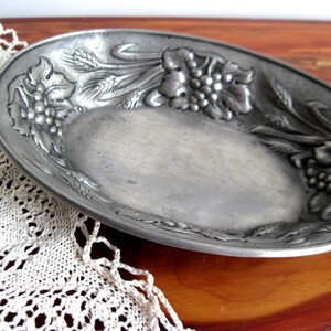 May include: An oval-shaped pewter dish featuring a raised relief design along the rim. The decorative pattern consists of clusters of grapes, grape leaves, and stalks of wheat. The metal has a matte, silver-gray finish with a smooth center.