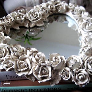 May include: A heart-shaped mirror framed by a dense arrangement of sculpted off-white roses. Each rose features distressed black edges, creating a weathered appearance. The reflective surface is smooth and clear, surrounded by the floral border.