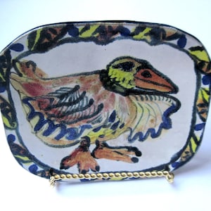 May include: An oval ceramic dish featuring a hand-painted illustration of a bird with orange, yellow, and blue plumage. The border is decorated with a repeating pattern of yellow and blue leaves. The dish rests on a small gold-toned metal stand.