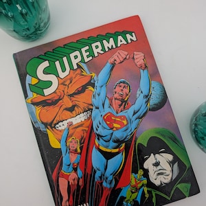 May include: A hardback Superman Official Annual 1983 comic book featuring a colourful cover illustration of Superman flying with his arms raised, alongside Supergirl, a large villainous face, and a hooded figure against a dark space background.