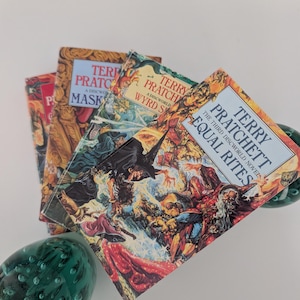 May include: A collection of three Terry Pratchett Discworld novels including Equal Rites, Wyrd Sisters, and Maskerade, featuring colorful illustrated covers with fantasy characters, positioned between two dark green glass decorative spheres.