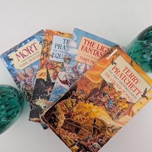 May include: Four paperback books by Terry Pratchett arranged in a fan shape. The titles include Mort, Equal Rites, The Light Fantastic, and The Colour of Magic. Each cover features colorful, detailed fantasy illustrations and bold, printed text.