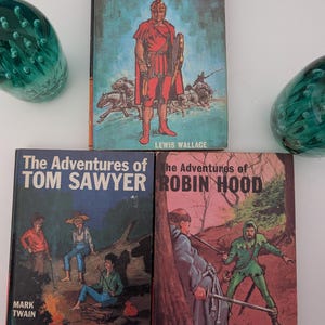 Op de afbeelding: Drie gebonden boeken met geïllustreerde covers: Ben Hur van Lewis Wallace met een Romeinse soldaat, De Avonturen van Tom Sawyer van Mark Twain met drie kinderen bij een kampvuur, en De Avonturen van Robin Hood met een bosscène.