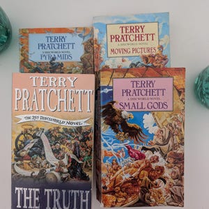 May include: Four paperback books by Terry Pratchett from the Discworld series. Titles include The Truth, Small Gods, Moving Pictures, and Pyramids. Each cover features detailed, colourful fantasy illustrations and bold text displaying the author's name.