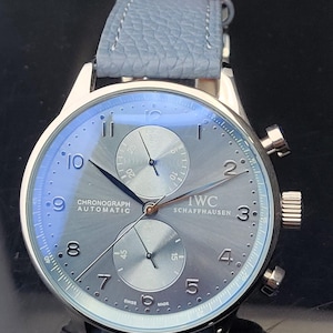 Puede incluir: Un reloj cron&oacute;grafo IWC Schaffhausen plateado con una correa de cuero azul claro. La esfera del reloj es gris con manecillas y n&uacute;meros negros. El reloj tiene dos subesferas y el texto "CHRONOGRAPH AUTOMATIC" es visible.