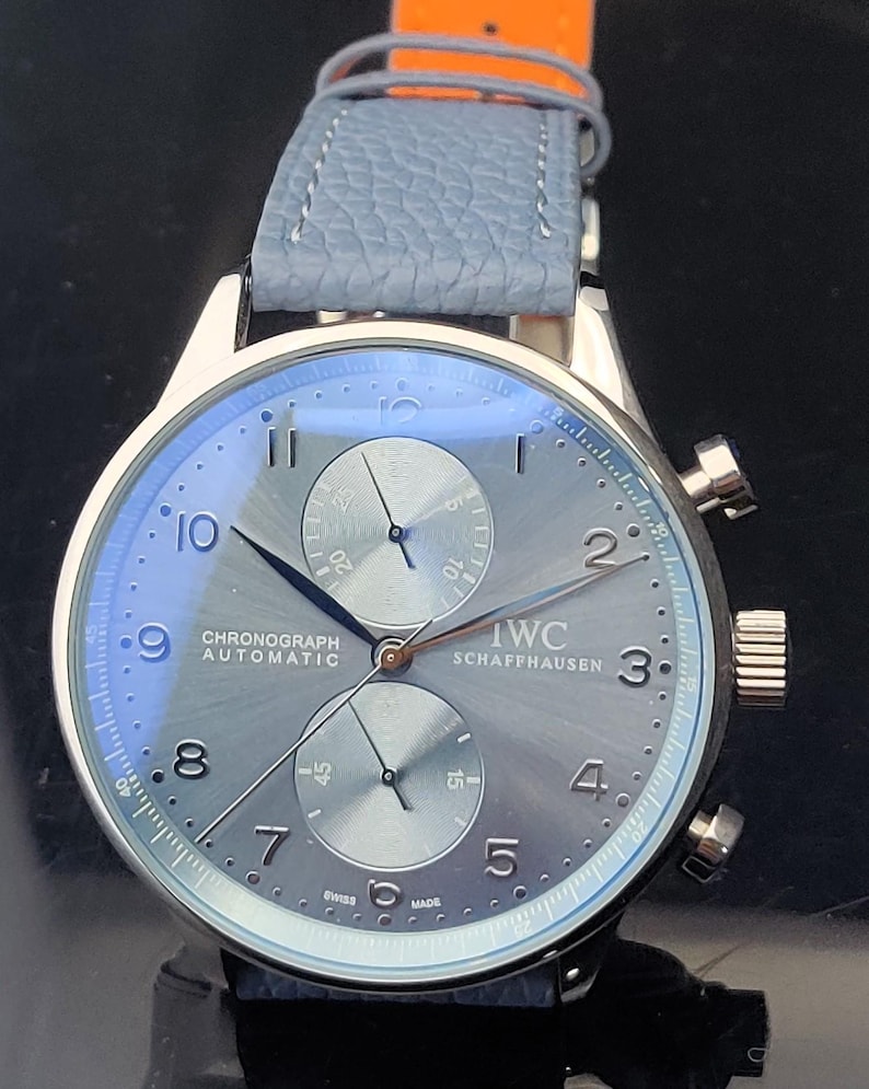 Puede incluir: Un reloj cron&oacute;grafo IWC Schaffhausen plateado con una correa de cuero azul claro. La esfera del reloj es gris con manecillas y n&uacute;meros negros. El reloj tiene dos subesferas y el texto "CHRONOGRAPH AUTOMATIC" es visible.