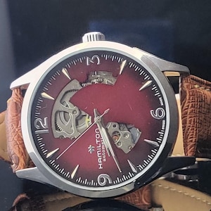 Op de afbeelding: Een zilveren Hamilton automatisch horloge met een bordeauxrode wijzerplaat en een bruine leren band. De wijzerplaat toont zichtbare interne mechanismen en de tekst "Hamilton automatic". De band is van getextureerd leer.