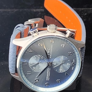 Puede incluir: Un reloj cron&oacute;grafo IWC Schaffhausen plateado con esfera gris y correa de cuero azul. El reloj tiene una esfera redonda con m&uacute;ltiples subesferas y el texto "CHRONOGRAPH AUTOMATIC". La correa tiene un interior naranja.