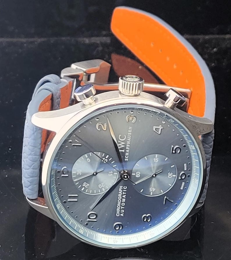 Puede incluir: Un reloj cron&oacute;grafo IWC Schaffhausen plateado con esfera gris y correa de cuero azul. El reloj tiene una esfera redonda con m&uacute;ltiples subesferas y el texto "CHRONOGRAPH AUTOMATIC". La correa tiene un interior naranja.
