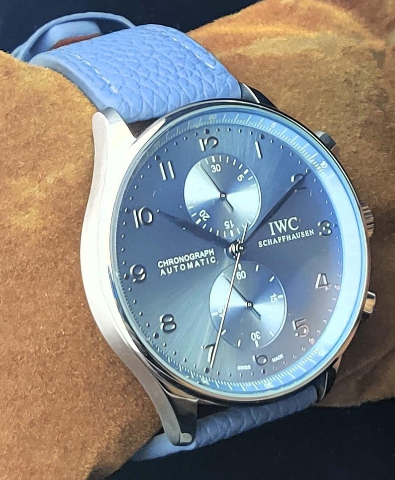 Puede incluir: Un reloj cron&oacute;grafo IWC Schaffhausen plateado con esfera azul y correa de cuero azul claro. La esfera del reloj muestra el texto "CHRONOGRAPH AUTOMATIC" y tiene dos subesferas. El reloj est&aacute; sobre un fondo marr&oacute;n.