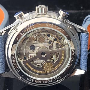 Puede incluir: Primer plano de un reloj cron&oacute;grafo plateado con una parte trasera transparente, que muestra los intrincados mecanismos internos. El reloj tiene una correa de cuero azul con detalles en naranja. Se ve el texto "IWC PORTUGIESER" y "CHRONOGRAPH".