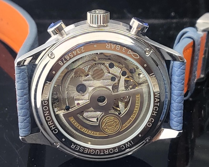 Puede incluir: Primer plano de un reloj cron&oacute;grafo plateado con una parte trasera transparente, que muestra los intrincados mecanismos internos. El reloj tiene una correa de cuero azul con detalles en naranja. Se ve el texto "IWC PORTUGIESER" y "CHRONOGRAPH".