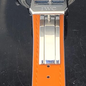 Puede incluir: Primer plano de un reloj de lujo con una correa de cuero azul claro y una parte inferior de cuero naranja. El reloj presenta un cierre plateado con el logotipo de IWC. La esfera del reloj es visible en el fondo.