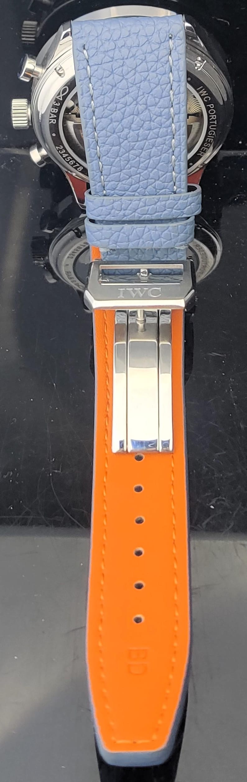 Puede incluir: Primer plano de un reloj de lujo con una correa de cuero azul claro y una parte inferior de cuero naranja. El reloj presenta un cierre plateado con el logotipo de IWC. La esfera del reloj es visible en el fondo.