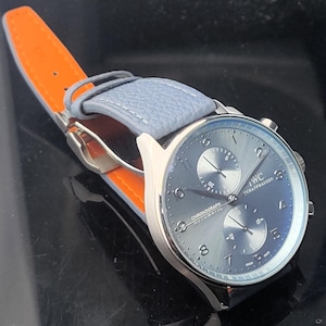 Puede incluir: Un reloj de pulsera plateado con esfera azul y correa de cuero azul claro. El reloj presenta dos subesferas y el texto "IWC SCHAFFHAUSEN CHRONOGRAPH AUTOMATIC". La correa est&aacute; sujeta a un respaldo de cuero naranja.