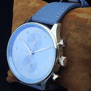 Puede incluir: Primer plano de un reloj de pulsera con esfera azul claro y correa de cuero a juego. El reloj tiene agujas plateadas y una caja plateada. La esfera muestra los n&uacute;meros del 1 al 12. El reloj est&aacute; sobre una superficie marr&oacute;n.