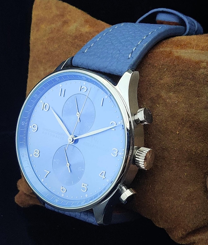 Puede incluir: Primer plano de un reloj de pulsera con esfera azul claro y correa de cuero a juego. El reloj tiene agujas plateadas y una caja plateada. La esfera muestra los n&uacute;meros del 1 al 12. El reloj est&aacute; sobre una superficie marr&oacute;n.
