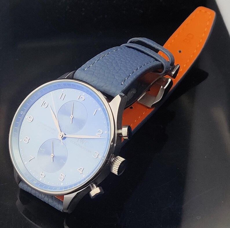 Puede incluir: Un reloj de lujo con esfera azul claro y correa de cuero azul. El reloj tiene una caja plateada. La correa est&aacute; sujeta a un respaldo de cuero naranja. El reloj est&aacute; en &aacute;ngulo, mostrando los detalles de la esfera y la correa.