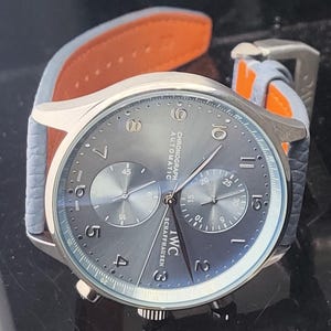 Puede incluir: Un reloj cron&oacute;grafo plateado con esfera gris y correa de cuero azul claro con forro naranja. La esfera del reloj muestra la marca "IWC SCHAFFHAUSEN" y el texto "CHRONOGRAPH AUTOMATIC". El reloj tiene dos subesferas y una visualizaci&oacute;n de la fecha.