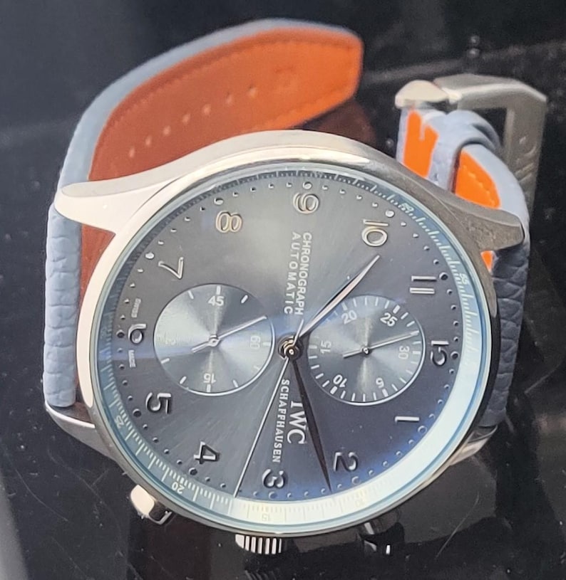 Puede incluir: Un reloj cron&oacute;grafo plateado con esfera gris y correa de cuero azul claro con forro naranja. La esfera del reloj muestra la marca "IWC SCHAFFHAUSEN" y el texto "CHRONOGRAPH AUTOMATIC". El reloj tiene dos subesferas y una visualizaci&oacute;n de la fecha.