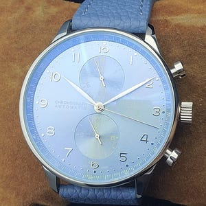 Puede incluir: Un reloj cron&oacute;grafo plateado con esfera azul claro y correa a juego. El reloj tiene una esfera redonda con manecillas y n&uacute;meros plateados, y dos esferas m&aacute;s peque&ntilde;as. El texto "CHRONOGRAPH AUTOMATIC" es visible.